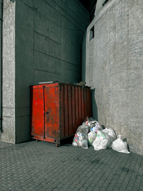 A large pile of black plastic rubbish bags filled with household waste is stacked outside a building, with some bottles and discarded packaging visible on top. Behind the bags, there is a tiled wall w