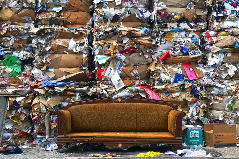 A red leather sofa is shown fallen onto the pavement outside a property, leaning against a stone wall, with the backrest resting on the wall and the seat facing downward. The sofa appears to be partia