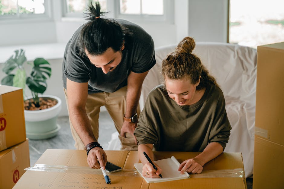 A person's hand holding a black pen and filling out a paper on a clipboard, which contains a moving or relocation checklist. The clipboard rests on a flat surface near a window or in a well-lit room,