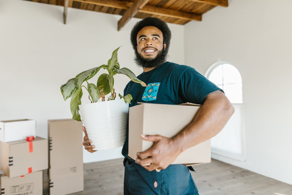 Three individuals standing outside a building, smiling and taking a selfie together while holding cardboard boxes with packing labels. The person on the left is wearing a maroon t-shirt, the one in th