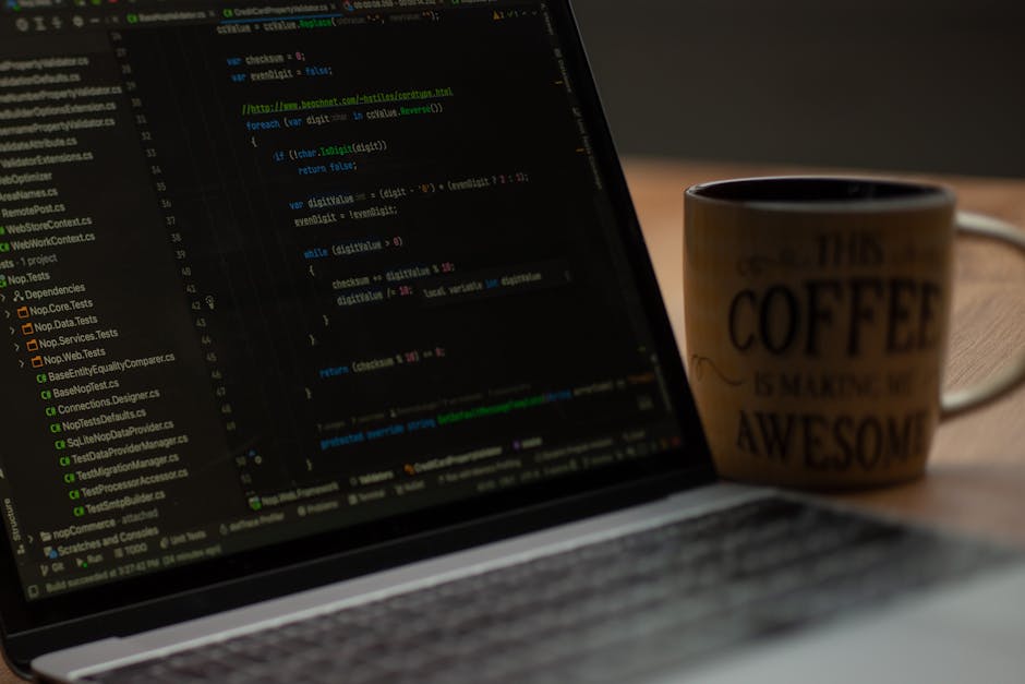 A laptop on a wooden desk displaying lines of code on a programming editor, with a ceramic coffee mug positioned to the right of the laptop. The mug features text that reads 'This Coffee is Making Me 